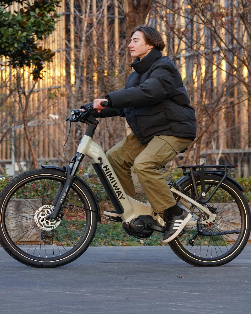 Junge Frau mit mittellangem Haar fährt entspannt auf einem hellen Himiway E-Bike durch einen ruhigen Parkweg, schwarze Steppjacke und olivgrüne Hose, weiches Licht am Morgen