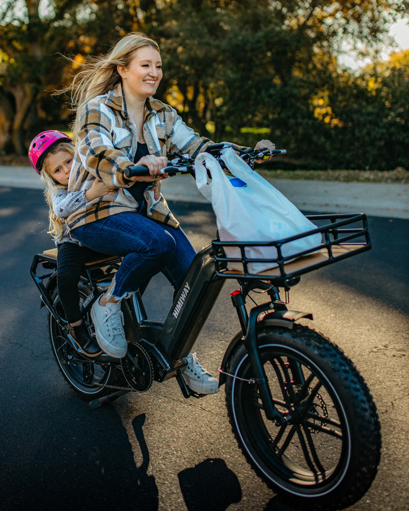 Junge Mutter mit langen blonden Haaren fährt entspannt mit ihrem Kind auf einem Himiway Cargo E-Bike einen sonnigen Weg entlang, das Kind mit rosa Helm lacht fröhlich, grüne Bäume im Hintergrund