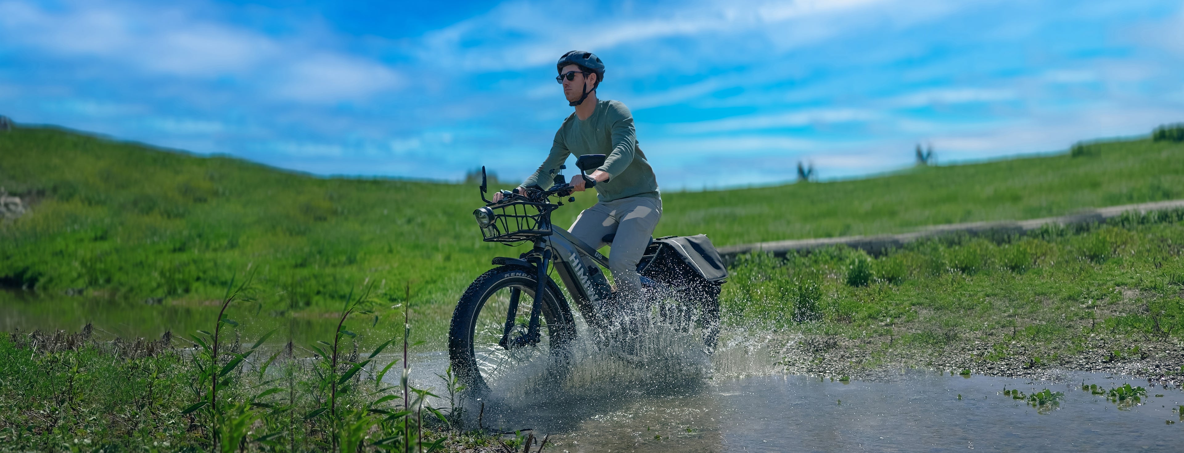 E-Bike-Fahrer überquert mit Schwung eine Wasserpfütze auf einem robusten Himiway, Sonnenlicht spiegelt sich auf spritzendem Wasser, grüne Felder und blauer Himmel im Hintergrundslider_item_NX7h4F