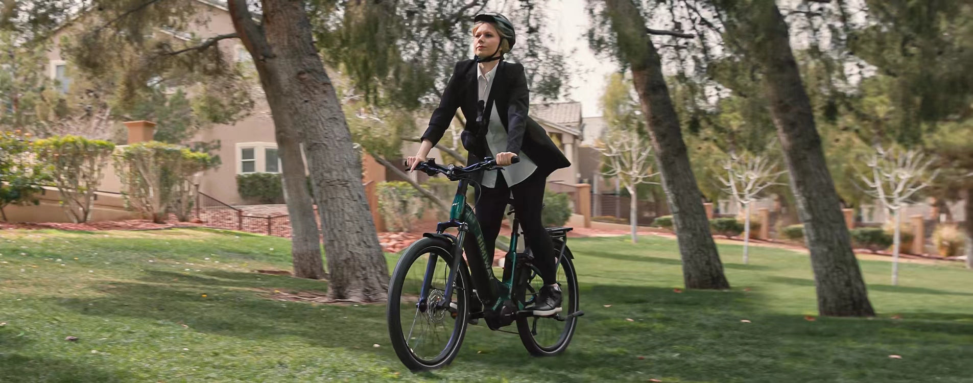 Stilvolle Frau im schwarzen Blazer fährt entspannt mit einem Himiway E-Bike durch einen sonnigen Park, grüne Bäume und Wohnhäuser im Hintergrundslider_item_8GM97q